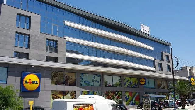 Το ολοκληρωμένο κτίριο γραφείων: γυάλινο κέλυφος με λευκές οριζόντιες ταινίες στους ορόφους και υπερμαρκετ στο ισόγειο, με τον δρόμο και τα αυτοκίνητα μπροστά.